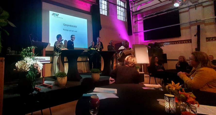Beeld van gesprekstafel 'Rondje uit het veld'. Drie mensen staan bij het presentatiescherm, in de zaal zitten mensen te luisteren