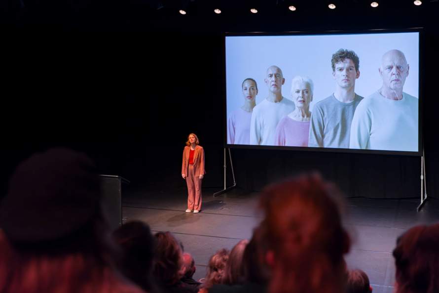De arts staat op het podium voor een schemr met mensen waarmee ze tijdens haar ziekte te maken krijgt.